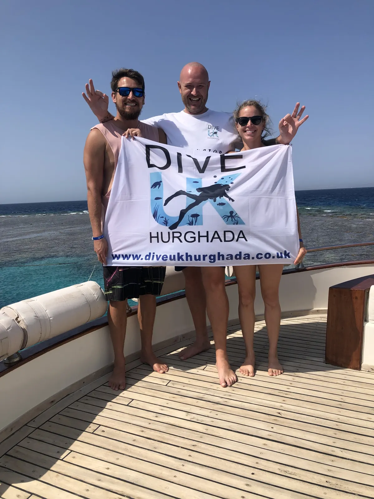 Three people holding a Dive UK Hurghada flag on a boat