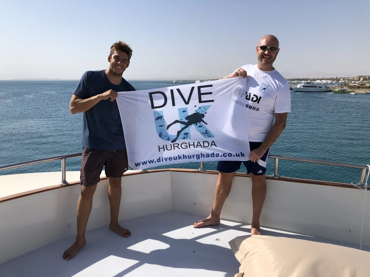 An instructor and guest holding a Dive UK Hurghada flag overlooking the sea
