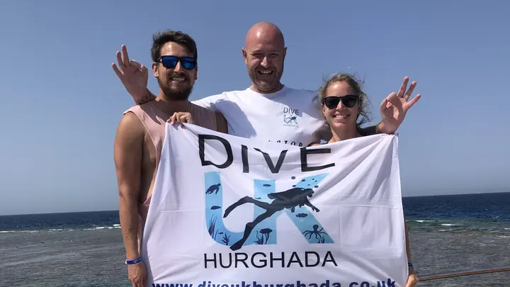 Three people holding a Dive UK Hurghada flag beside the sea