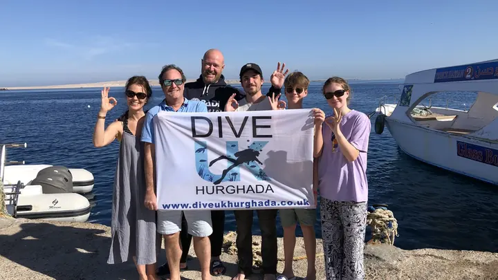 A group holding a Dive UK Hurghada flag beside the sea