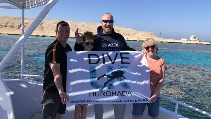 Four people holding a Dive UK Hurghada flag on a boat