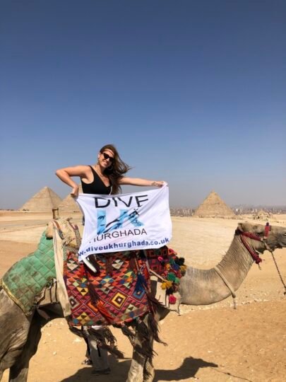 Guest holding the Dive UK Hurghada flag on a camel with the pyramids behind her