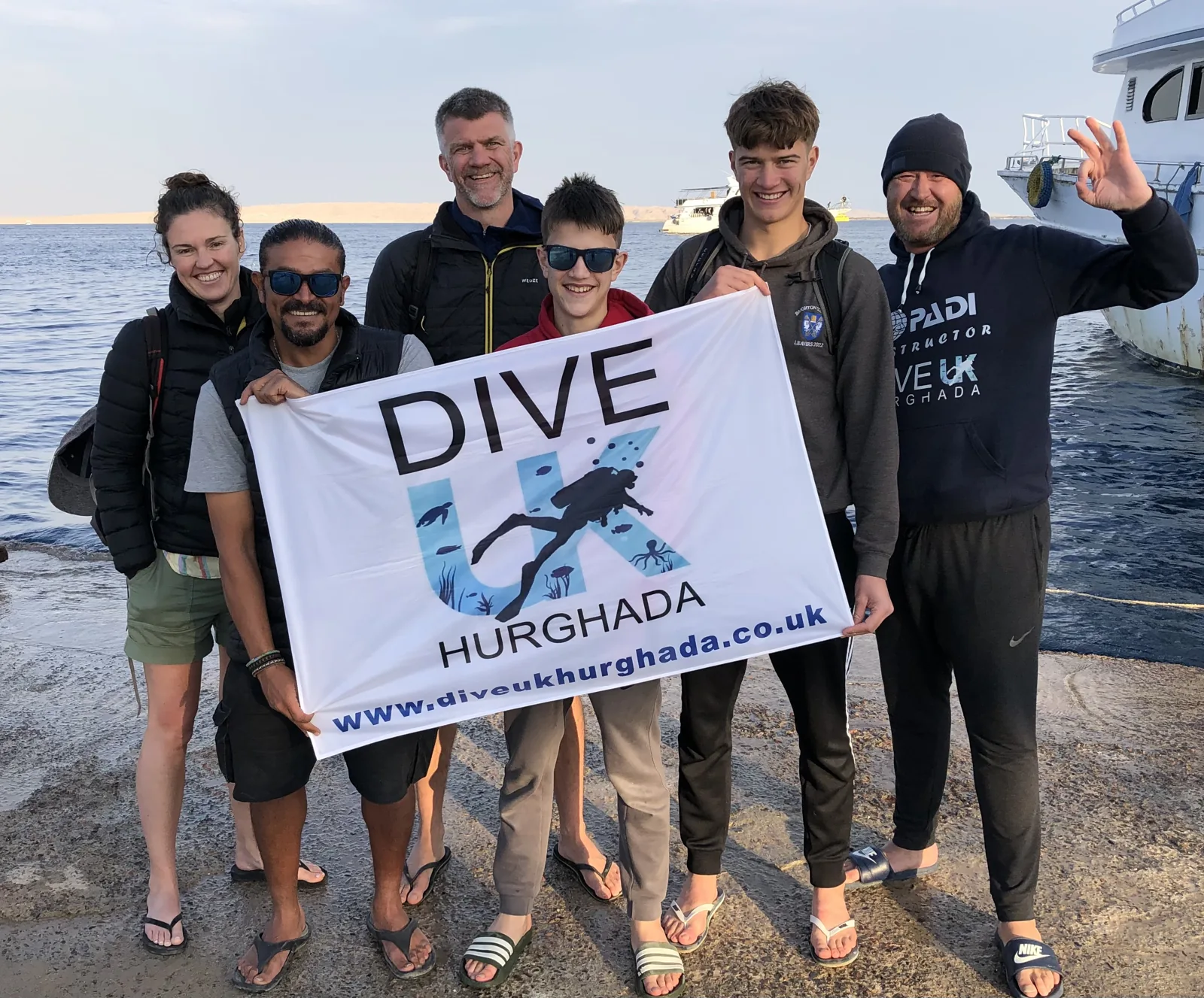 Guests and instructors holding a Dive UK Hurghada banner beside the Red Sea